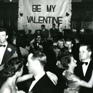 A black and white photo of students dancing in the quad dining hall in 1950. Students are all dressed formally and dancing in couples. A large sign hanging on the wall reads BE MY VALENTINE, and a small band performs in front of it.