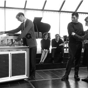 A black and white photo of five students in the forum in 1965. One student is selecting a song on the jukebox, while two students stand two his right, pausing their slow dancing to watch him select a song. Two more students sit on the steps behind the dance floor, watching the couple.