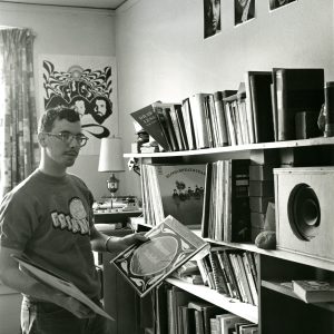 A black and white photo of a male student standing in front of his record collection. He is wearing a Grinnell T shirt and holding a Telemann Pachebel record out for the camera. The walls have posters of rock bands.