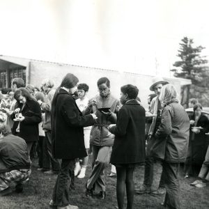 A black and white photo of students standing gathered in groups, talking outside of Burling in 1971. They are wearing coats and eating out of various types of dish ware.