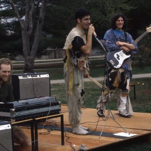 A color photo of three male students performing on the commencement stage in 1976. From left to right, a student with long hair is sitting behind a mixing board, a student draped in multi-colored rags and not wearing shoes is holding a microphone, and another student draped in rags holds a guitar.