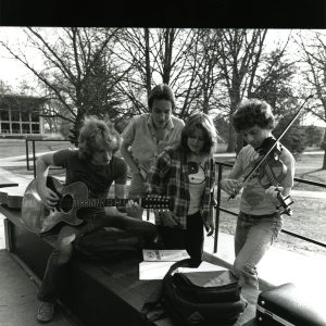 A black and white photo of four students sitting in front of the forum in 1983, looking down at music in front of them. Two students play instruments, a guitar and a violin.