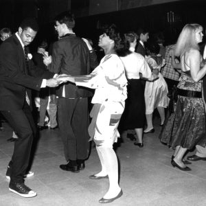A candid photo of a waltz at Grinnell in 1986. Two couples are in focus, both in the midst of dancing with their partners. Other students and couples fill the background of the image. Everyone is wearing semi-formal wear, such as suits and mid-length dresses.