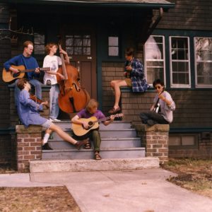Six students sit sprawled on the front steps of Russian House, in a candid photo during a musical performance. Three students sit in the back, from left to right playing a guitar, a Cello, and what appears to be a Ukulele. The three students in the front, from left to right, are playing a violin, guitar, and banjo.