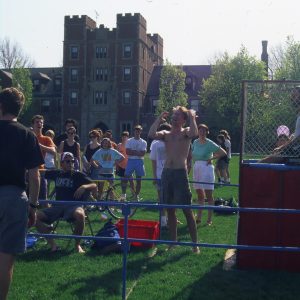 A group of students from 1992 stand on Mac Field, surrounding a man sitting in a dunk tank. The tank is fenced off, and the people are looking off camera, presumably at another student throwing balls at the tank.